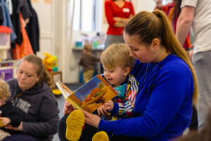 A woman in a blue jumper reading a book to a young boy who is smiling, in a playgroup setting.