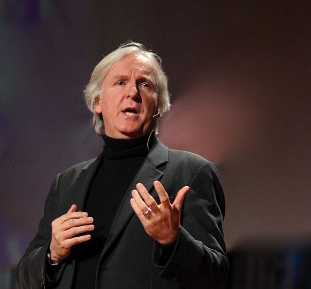 A white man with mid-length grey hair wearing a suit and turtleneck, talking emphatically on stage