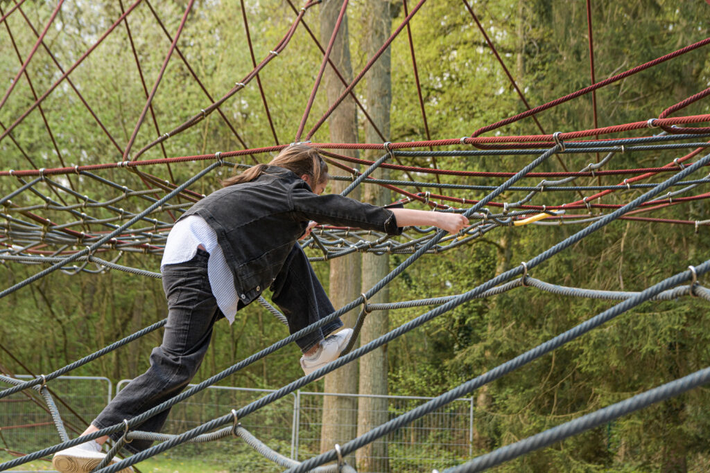 Teenage girl climbing on spider web with expression in public park.