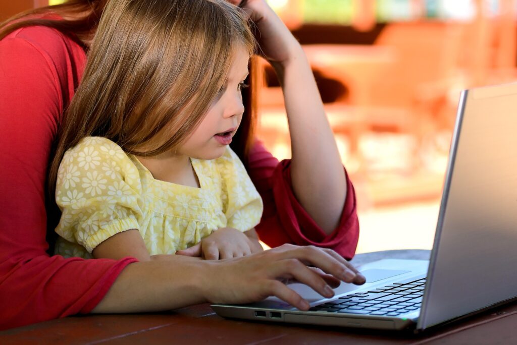 A young child sat on her mother's lap working on a task on a laptop.