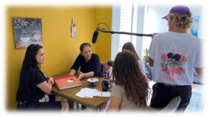 People talking at a table with a microphone, being filmed
