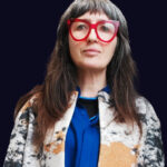 A portrait image of Cristina Cerulli wearing an electric blue shirt and red glasses